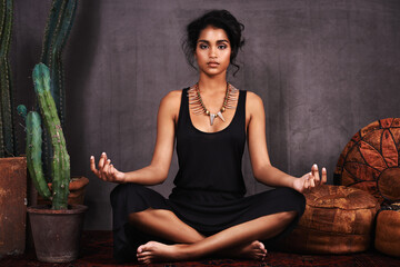 Meditating magic. Studio portrait of a beautiful young woman sitting in the lotus position beside a cactus and cushions.