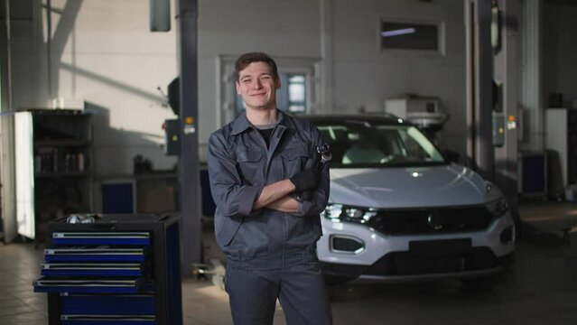 car service, a young male mechanic in uniform with tools in his hands stands on road of a vehicle for diagnosing breakdowns, smiling and looking at camera