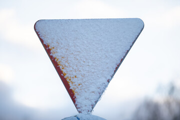 A snowy triangular road sign, give way to priority against a blurred sky.