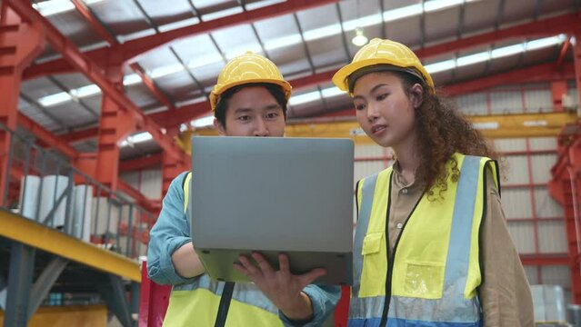 Two Asian worker man and woman team working wear safety uniform with hardhat meeting with laptop in metal sheet factory industry workshop, Engineering talk and showing data in notebook computer