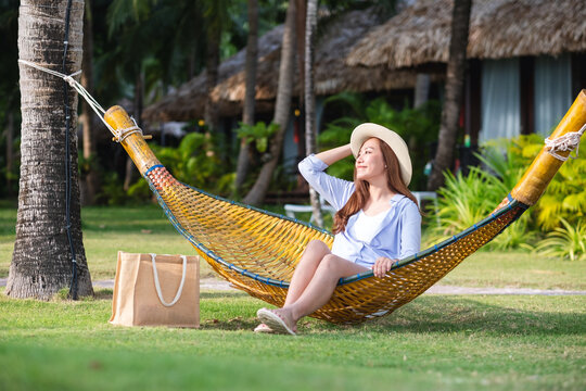 Portrait image of a beautiful young asian woman with hat sitting on bamboo hammock in a tropical garden