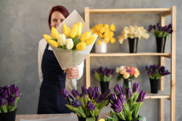 concentrated young female florist in a ftruk works in a flower shop. Stands near a showcase with flowers, holding a bouquet of yellow tulips in his hands.