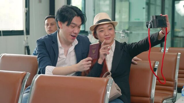 Wait boarding flight. Asian beautiful young loving couple smiling making selfie by their mirrorless camera and holding passports tickets while waiting for boarding in terminal hall airport