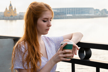 Thoughtful young woman standing at fence near the river while holding paper cup with takeaway coffee 