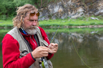 Mature fly fishing fisherman in polarizing glasses tying fly of fishing hook on fishing rod