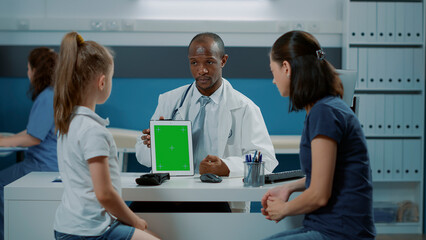 Fototapeta premium African american medic holding green screen on digital tablet, sitting with parent and kid. Doctor showing device with isolated mockup template, copy space background and blank chroma key.