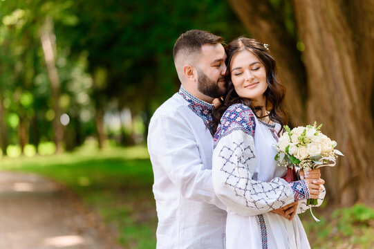 Side view of bearded man, which wearing in embroidery shirt, standing behide of girl and hugging her waist. Attractive bride in long white gown with pattern detail, holding bouquet of flowers