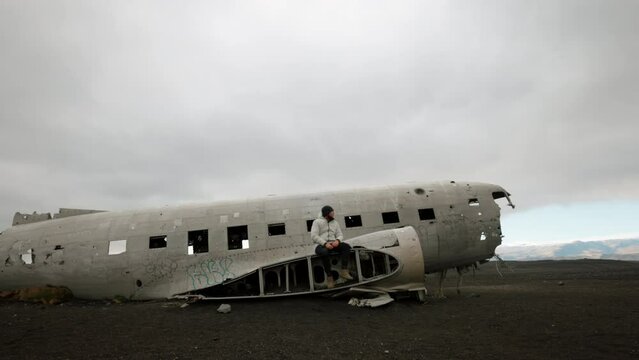 Man in white lonely sad sitting on old crashed broken wreck Icelandic plain dc-3 landmark in Iceland. High quality 4k footage in slow motion. Apocalyptic catastrophe lost view