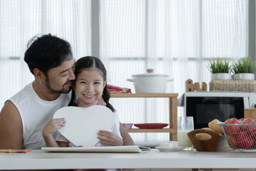 Happy Caucasian little daughter smiling holding heart shaped card and giving to her Asian young father after finished drawing and making gift for dad on fathers day at home kitchen with love