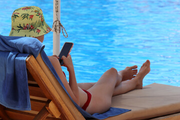 Slim girl in bikini lying with smartphone in hands in deck chair near swimming pool in shadow of...