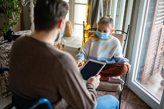 Father with disability in wheelchair using tablet at home with child while wearing masks.