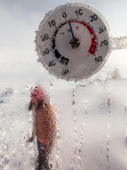 A round plastic thermometer measures the temperature of the air outside the fogged window of a country house. Silhouette of a woman weared in warm winter clothes in the background