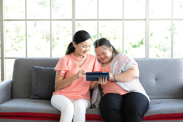 Obraz premium mother using tablet computer with a girl down syndrome or her daughter, smiling and enjoying on sofa