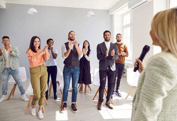 Fototapeta premium Seminars and conferences. Happy grateful people at business training applaud female speaker who shared with them his experience and knowledge. Young entrepreneurs applaud unknown woman with microphone