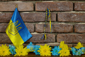 National flag of Ukraine with the ukrainian coat of arms, blue and yellow chrysanthemums, yellow and blue ribbons are against a brick wall 