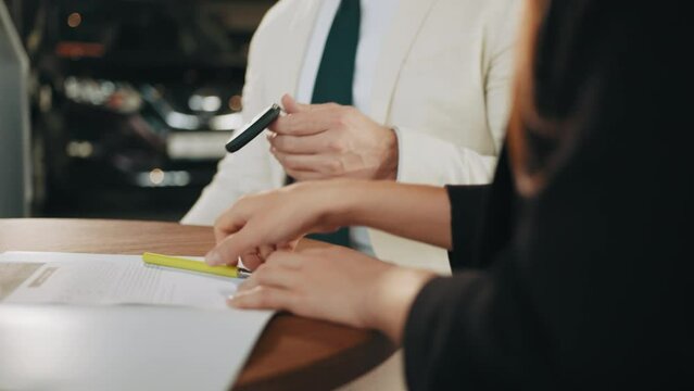 Multi ethnic couple signing documents about car purchasing and receiving keys from salesman. Happy customers making successful deal at modern showroom. Car dealer giving keys from new auto.