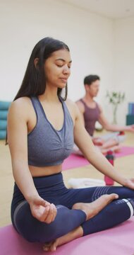 Vertical video of biracial woman with eyes closed meditating with legs crossed in yoga class
