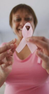 Vertical video of smiling senior african american woman holding pink breast cancer ribbon to camera