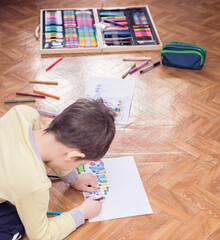 The child boy sits on the floor and draws a picture with colored markers on white sheet. Leisure activity at home