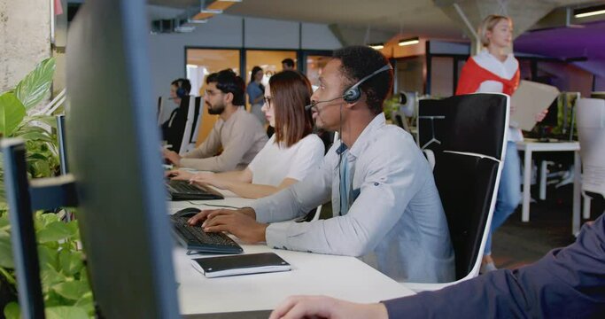 African American man in headset working at computer and talking with client in office. Mixed-races support team of company. Workers of call center. Chatting with customers. Selling online.