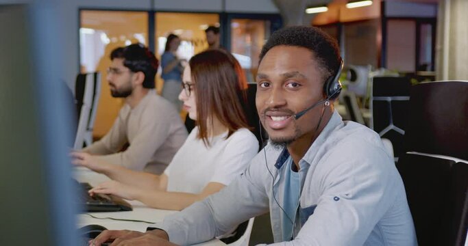 African American young man in headset talking with client at computer. Call center concept. Portrait of happy male support worker looking at camera and smiling. Job in sales. Selling by phone.