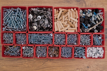 Various size and shape of screws, bolts, wooden pegs selection in red plastic tray box on a wooden background. Assembly kit for furniture. Top view.