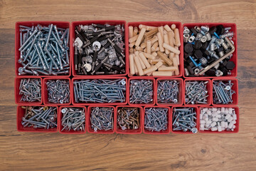 Various size and shape of screws, bolts, wooden pegs selection in red plastic tray box on a wooden background. Assembly kit for furniture. Top view.