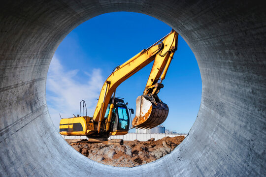 A powerful caterpillar excavator digs the ground against the blue sky. Earthworks with heavy equipment at the construction site. View through a large concrete pipe.