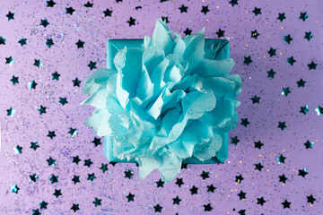 A gift wrapped in blue eco kraft paper with a lush bow on a lilac background with small confetti in the form of stars.The holiday, surprise, birthday.