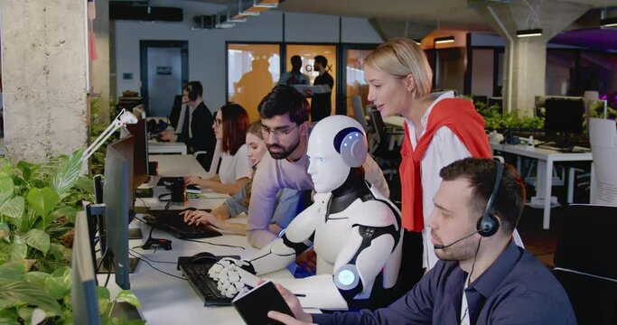 Happy multietnic people standing and looking at humanoid working in office. Robot typing on keyboard at call center. Futuristic support service. Men and women cheering up for android work.