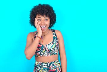 young woman with afro hairstyle in sportswear against blue background excited looking to the side hand on face. Advertisement and amazement concept.