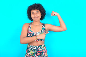 Smiling young woman with afro hairstyle in sportswear against blue background raises hand to show...