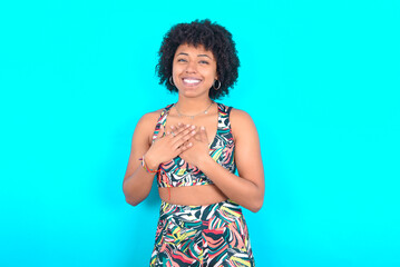 Honest young woman with afro hairstyle in sportswear against blue background keeps hands on chest,...