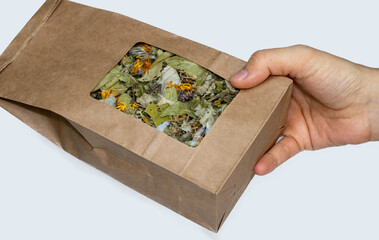 brown craft recycled paper with window with natural tea leaves, harvest from garden. woman's hand is holding big paper eco pack isolated on grey background. healthy tea components. Organic tea brand, 