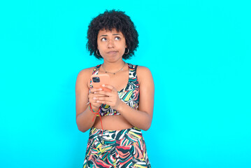 Portrait of young woman with afro hairstyle in sportswear against blue background with dreamy look, thinking while holding smartphone. Tries to write up a message.