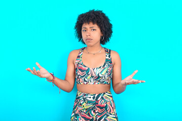 young woman with afro hairstyle in sportswear against blue background looks uncertain shrugs...