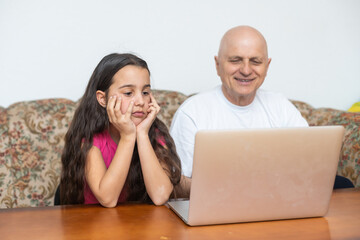 Adorable little girl hugging happy grandfather using laptop at home.