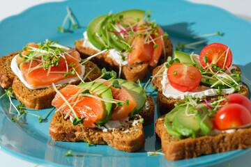 closeup toast with salmon, trout, cherry tomatoes, avocado and microgreens on blue plate, open sandwich, healthy breakfast snack