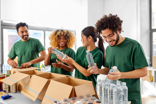 charity, food donation and volunteering concept - international group of happy smiling volunteers packing bottles of water in boxes at distribution or refugee assistance center