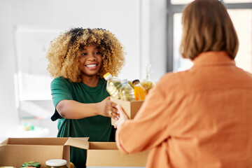 charity, donation and volunteering concept - happy smiling female volunteer and woman taking box of food at distribution or refugee assistance center
