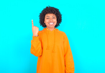 Obraz na płótnie Canvas young woman with afro hairstyle wearing orange hoodie against blue background smiling and looking friendly, showing number one or first with hand forward, counting down