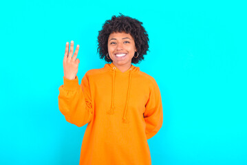 Obraz na płótnie Canvas young woman with afro hairstyle wearing orange hoodie against blue background smiling and looking friendly, showing number three or third with hand forward, counting down