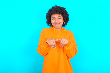 Obraz na płótnie Canvas young woman with afro hairstyle wearing orange hoodie against blue background makes bunny paws and looks with innocent expression plays with her little kid