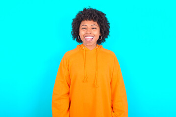 Obraz na płótnie Canvas Funny young woman with afro hairstyle wearing orange hoodie against blue background makes grimace and crosses eyes plays fool has fun alone sticks out tongue.