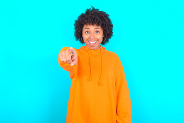 Obraz na płótnie Canvas Excited positive young woman with afro hairstyle wearing orange hoodie against blue background points index finger directly at you, sees something very funny. Wow, amazing