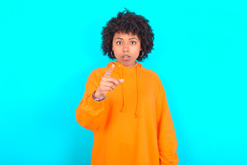 Obraz na płótnie Canvas Shocked young woman with afro hairstyle wearing orange hoodie against blue background points at you with stunned expression