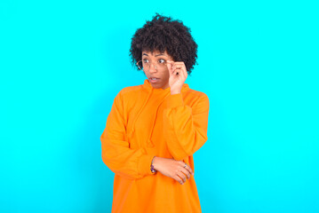 Obraz na płótnie Canvas Astonished young woman with afro hairstyle wearing orange hoodie against blue background looks aside surprisingly with opened mouth.