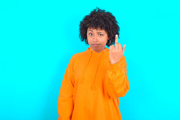 young woman with afro hairstyle wearing orange hoodie against blue background shows middle finger bad sign asks not to bother. Provocation and rude attitude.