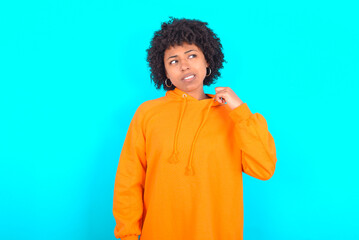 Obraz na płótnie Canvas young woman with afro hairstyle wearing orange hoodie against blue background stressed, anxious, tired and frustrated, pulling shirt neck, looking frustrated with problem