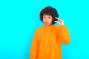 Obraz na płótnie Canvas young woman with afro hairstyle wearing orange hoodie against blue background purses lip and gestures with hand, shows something very little.